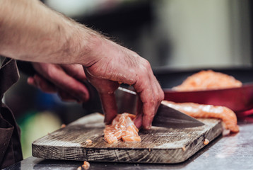 Male Chef Cutting Fresh Salmon on the Wooden Board with Blurred Pan in a Background