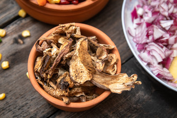 Dried boletus mushrooms in rustic ceramic bowl on old wooden table, and other mixture of vegetables and spices in the background. 