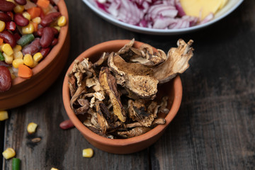 Dried boletus mushrooms in rustic ceramic bowl on old wooden table, and other mixture of vegetables and spices in the background. 