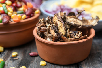 Dried boletus mushrooms in rustic ceramic bowl on old wooden table, and other mixture of vegetables and spices in the background. 