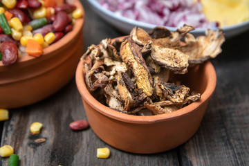 Dried boletus mushrooms in rustic ceramic bowl on old wooden table, and other mixture of vegetables and spices in the background. 