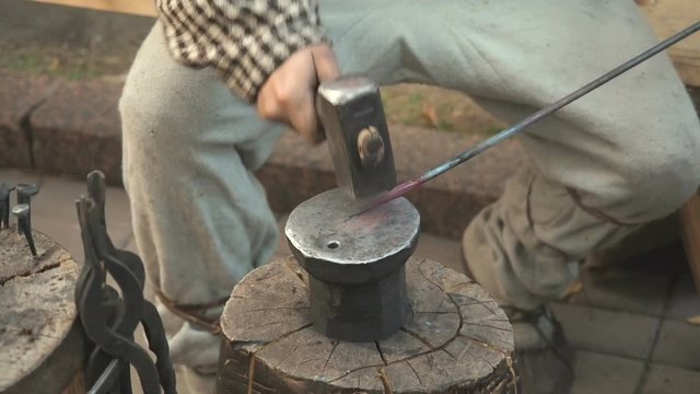 The blacksmith in the production process of metal products handmade on the openair workshop. Blacksmith strikes with a hammer on metal. Hands of a blacksmith with red-hot billet close up
