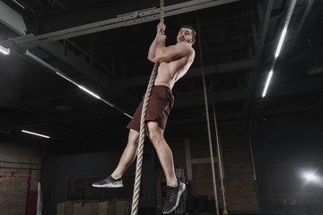 Young crossfit athlete climbing a rope at the gym. Man doing functional training. Workout exercises