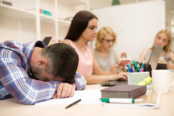 View of male student tired student sleeping on his books, during lecture at university. Group of students sitting at table, reading book on blur background in class.