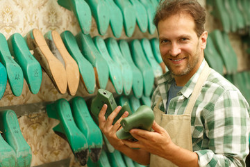 Man in checkered shirt and an apron standing next to shelves with details for sewing shoes and...