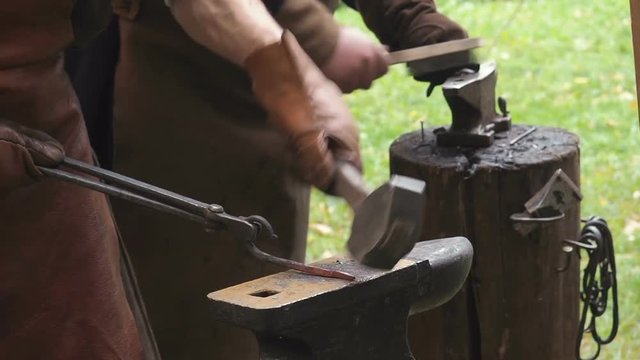 Two blacksmiths in the production process of metal products handmade on the openair workshop. One of blacksmiths strikes with a hammer on metal, another cleans the workpiece from the scale and soot