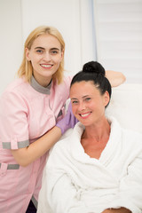 Portrait of young female cosmetologist and beautiful smiling patient during procedure in beauty parlor. Satisfied brunette in white robe sitting and professional doctor in uniform standing aside.