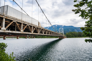Heavy Truck crossing a Suspesion Bridge Spanning a Large River in the Mountains on a Cloudy Summer Day