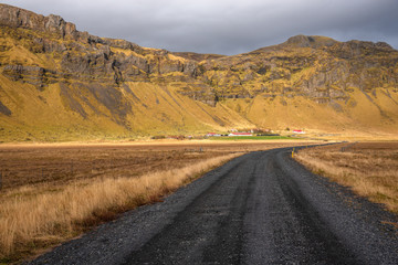 Fototapeta na wymiar Dirty Road Leading to a Farm at the Foot of a Mountain in the Countryside of Iceland under Autumnal Stormy Sky 