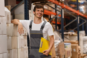 Cheerful worker wearing uniform and white t shirt, holding yellow clipboard. Handsome man smiling, standing and leaning on white boxes in warehouse. Concept of entrepot and commercial industry.