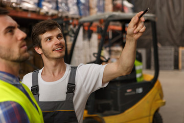Handsome worker smiling and showing something to his colleague. Cheerful man wearing uniform and white t shirt. Concept of transportation of goods, warehouse and entrepot.