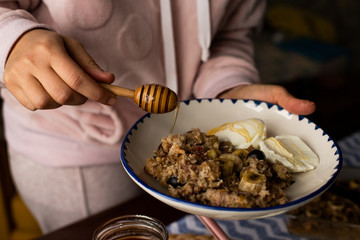 preperation of baked oatmeal with banana, blueberries, walnut and raisins. served with yoghurt and honey. dripping honey. putting yoghurt.