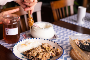 preperation of baked oatmeal with banana, blueberries, walnut and raisins. served with yoghurt and honey. dripping honey. putting yoghurt.