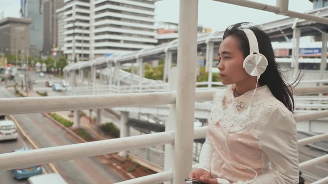 A sad Indonesian girl walks through the transition in the center of the city in white headphones and listens to music.