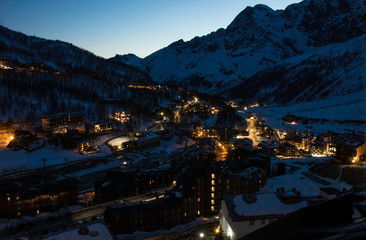European mountains landscape, Alps, Italy, mountain skiing resort, picturesque view with deep blue sky, high rock peaks on horizon, town village houses lights at night photo. Tourism, travel, resort