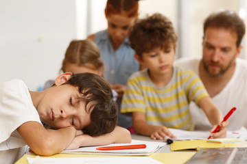 Fototapeta na wymiar Lazy little student in white shirt sleeping during lesson in classroom. Exhausted with boring study boy leaning on table while classmates writing with teacher on background.