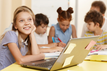 Portrait of smart pupil sitting at table, thinking ideas and looking away in classroom. Smiling little girl with classmates on background studying and using computer during lesson in school.