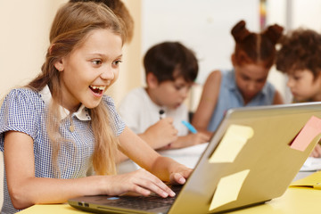 Curious pretty schoolgirl using laptop and typing in classroom. Smart girl studying and using...