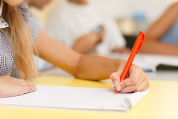 Closeup of hands of little schoolgirl writing with orange pen on notebook during lesson in classroom. Process of creating essay, learning new information and studying in modern school.