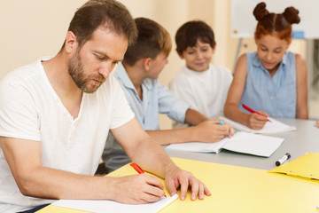Serious teacher in white shirt carefully checking works, sitting at table near little students, keeping pen and looking at notes in classroom. Specialist within field of education in process of work.