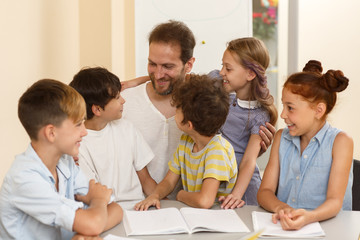 Positive professional male teacher with little cute students standing near table, smiling during lesson to each other. Smart children in process of studying in modern school.