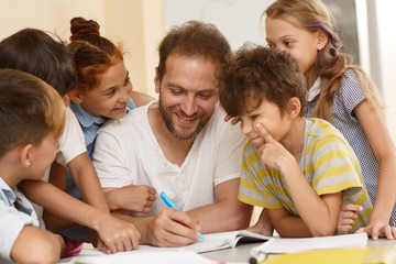 Positive male teacher in process of teaching little curious students in classroom. Smiling tutor sitting at table, writing and explaining material, smiling children standing around and listening.