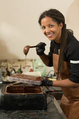 Side view of smiling woman confectioner pouring chocolate with scoop in baling forms. Afro female cook wearing uniform in process of preparing chocolate desserts at kitchen, looking at camera.