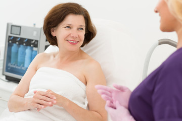 Obraz premium Woman having appointment with doctor cosmetologist. Female client lying on couch and talking with beautiful beautician in uniform and gloves. Concept of skin care and treatment.