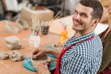 View from back of handsome man practising craft in workshop and enjoying working process. Smiling master in checkered shirt and red apron sculpting bowl from clay, looking at camera and posing.