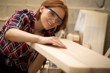 Female joiner holding lumber in hands, checking quality and looking at it.  Beautiful and positive craftswoman with ginger hair wearing safety glasses and checked shirt.