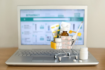 Shopping cart toy with medicaments in front of laptop screen with pharmacy web site on it. Pills, blister packs, medical bottles, thermometer set. Health care and internet shopping. - 251499602