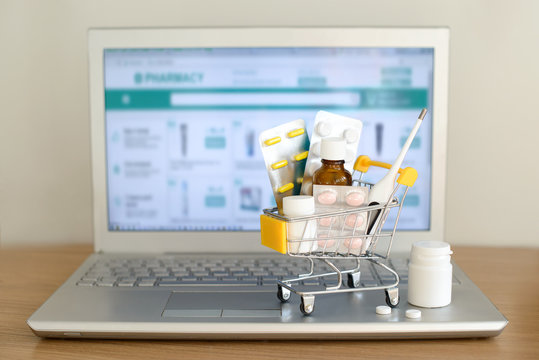 Shopping cart toy with medicaments in front of laptop screen with pharmacy web site on it. Pills, blister packs, medical bottles, thermometer set. Health care and internet shopping.
