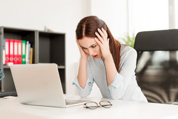 Business concept - confused young business woman sitting at her desk with the laptop in the office. Problems at work