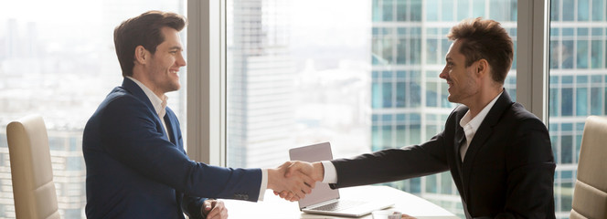 Horizontal image businessmen in suits handshaking sitting at office desk