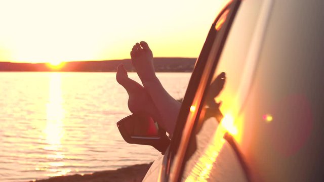 LENS FLARE, CLOSE UP: Happy girl kicking her feet to the beat of the music playing in her car. Beautiful sunset shines on the unrecognizable woman's feet while enjoying a relaxing evening by seaside.