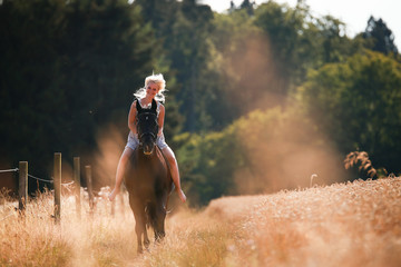 Young girl rides her horse, on a summer evening at sunset, in a summer dress past grain fields.