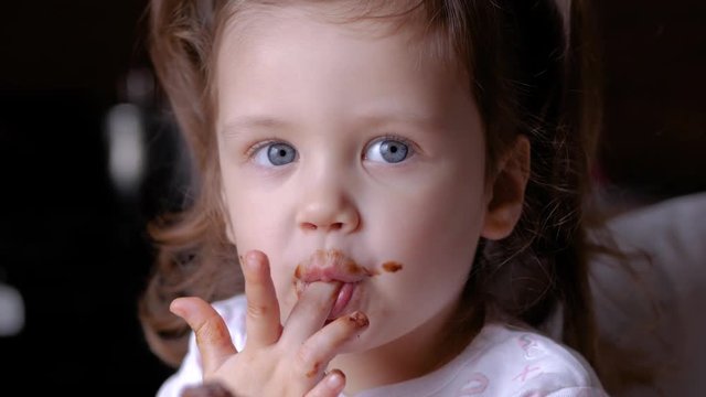Closeup view of funny dirty small girl after eating chocolate. Brown lips and teeth. Child licking fingers.