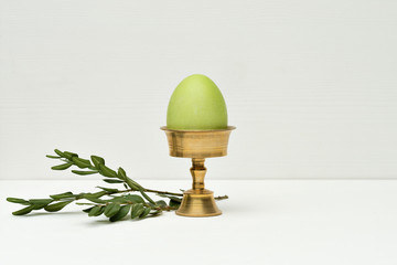 Green easter egg on stand and twig on white wooden background
