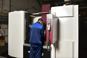 The worker in blue workwear, the operator of the CNC machine, oversees the process and controls it. A man works in a factory with CNC machine