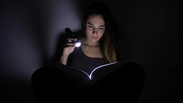 close-up portrait girl is reading a book with a flashlight in a dark room at night. 4k. Motion shooting