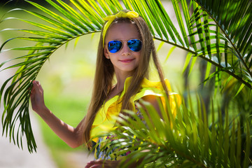 Beautiful girl with long hair in a yellow swimsuit and multi-colored sunglasses on the background of palm trees