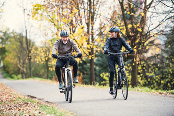 A senior couple with electrobikes cycling outdoors on a road in park in autumn.