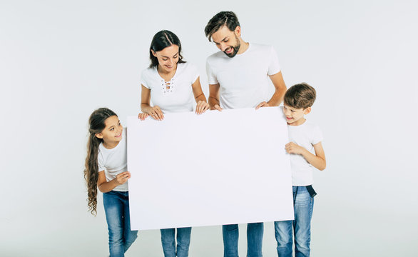 Beautiful Smiling Family In White T-shirts Hold In Hands The Big Blank Banner And Looking On Camera