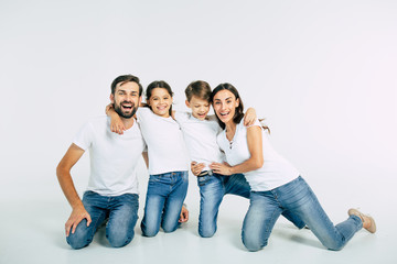 Relationship concept. Beautiful and happy smiling young family in white T-shirts are hugging and have a fun time together while sitting on the floor and looking on camera.