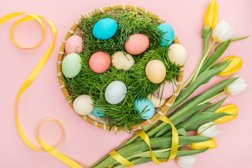 top view of easter eggs in wicker plate with grass isolated on pink with tulip flowers