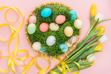 top view of easter eggs in wicker plate with grass isolated on pink with tulips