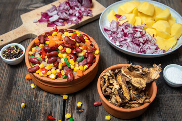 Mixture of vegetables, dried boletus mushrooms, sliced potato, red onion sliced and whole on a cutting board and spices, on old rustic wooden table.