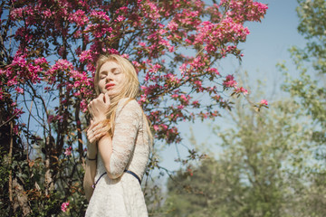 Beautiful young blonde woman in white lace dress enjoying blooming nature with closed eyes.