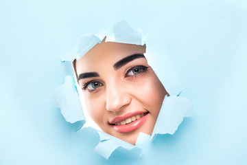 the face of a young beautiful girl with a bright make-up and puffy blue lips peers into a hole in...