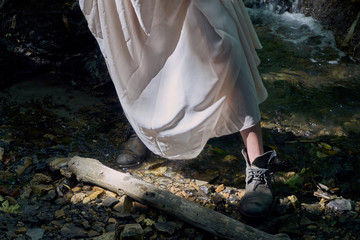 The legs of a young girl in a light summer dress and vintage shoes standing by a small stream on a clear sunny day in the forest. Through the thin dress the sun's rays shine
